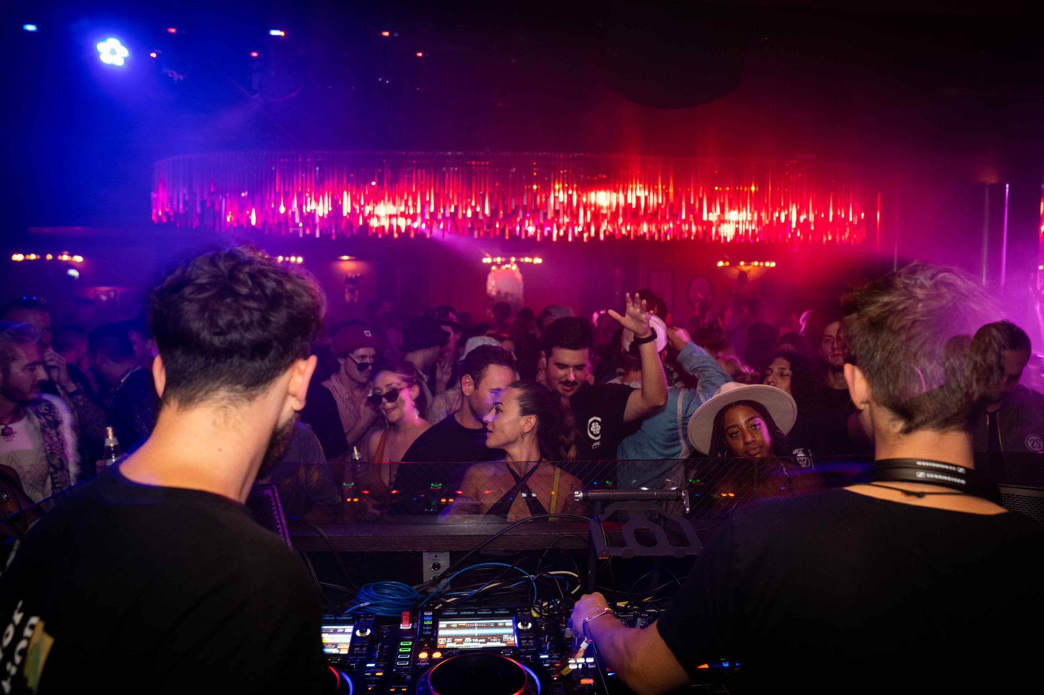 A dark, crowded nightclub or concert venue with a stage illuminated by bright red and blue lights, with silhouettes of people in the foreground and on the dance floor.