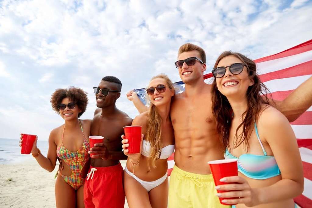 A group of diverse friends celebrating American Independence Day, holding red cups and smiling, with an American flag draped behind them. 