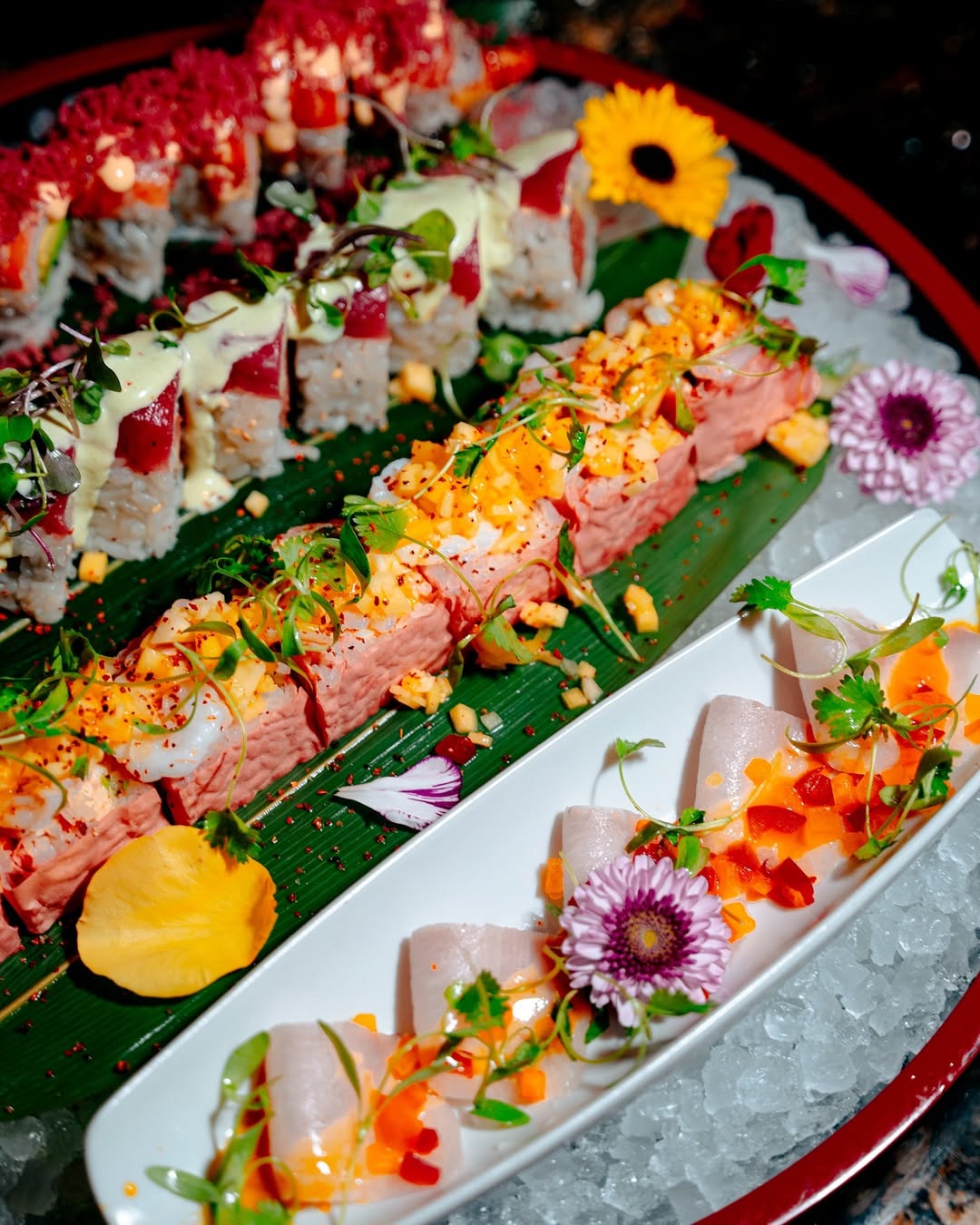A vibrant platter of various sushi rolls and sashimi, beautifully arranged on a dark red and black serving tray with ice and decorative flowers, including a yellow sunflower and purple chrysanthemums.