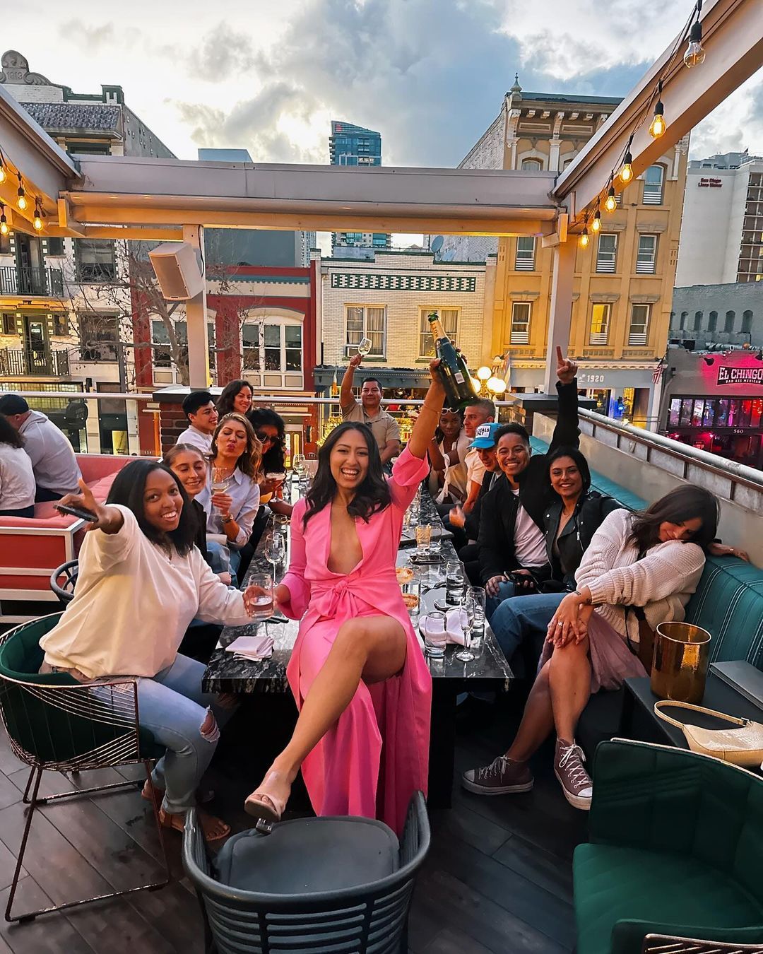 A group of diverse people enjoying a lively evening on the rooftop patio of Rustic Root in San Diego's Gaslamp Quarter, featuring a woman in a pink dress raising a bottle of champagne in celebration amidst a backdrop of city buildings and string lights