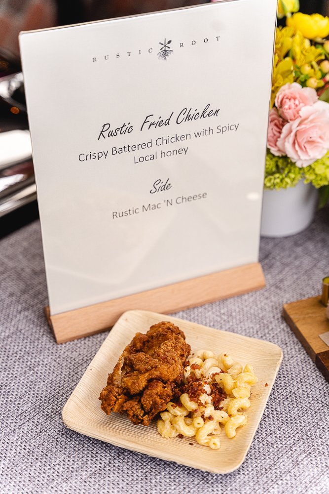 A plate of Rustic Fried Chicken with a side of Rustic Mac 'N Cheese is displayed on a wooden surface next to a white sign with the Rustic Root logo.