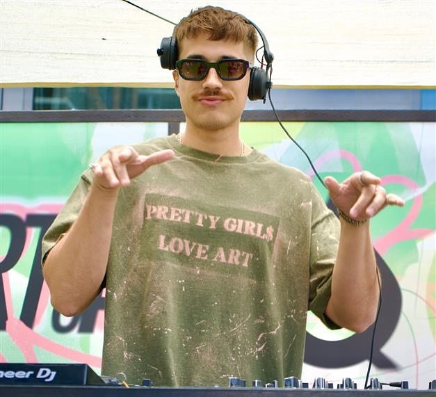 A young man with headphones and sunglasses, wearing an olive green t-shirt that says "PRETTY GIRLS LOVE ART," stands behind DJ equipment, gesturing with his hands.