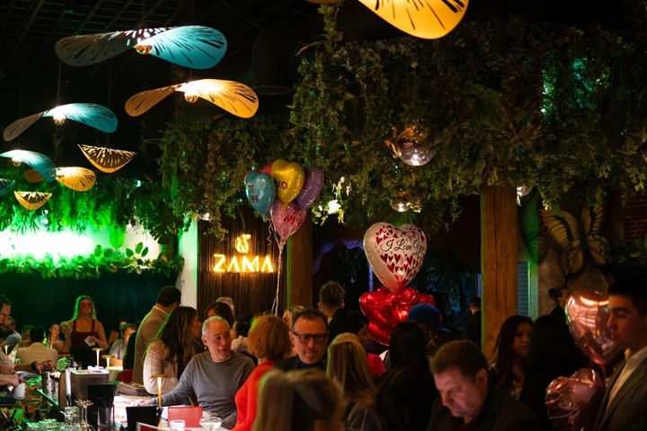 A lively interior view of Zama, a restaurant and bar, with numerous patrons gathered around tables and a bar area.
